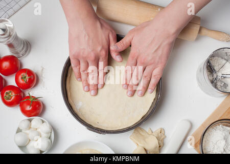 Male chef puts the rolled out dough in a pizza shape. On the white table are the tomatoes, mozzarella balls, olive oil and flour. Stock Photo