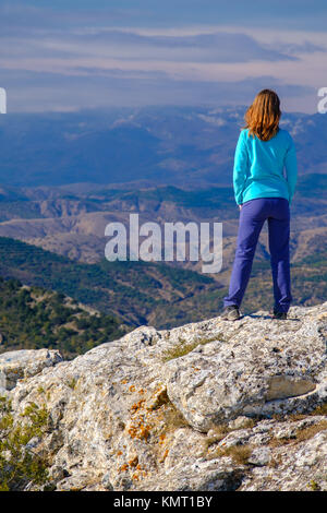 Athletic young woman standing on the rocky top of the mountain against the blue sky. Back to the camera Stock Photo