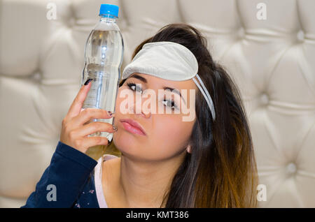 Drunk woman with a sleeping eye mask in her head and with a bottle of water pressing in her face using her other hand, hangover concept Stock Photo