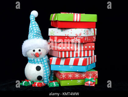 Small snowman standing next to a pile of colorful boxes Christmas Presents stacked high above his head. Isolated on black background Stock Photo