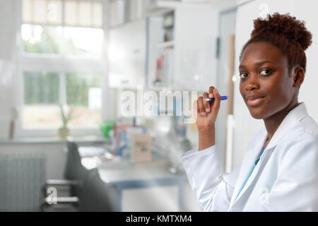 African scientist, medical or or graduate student. Bright, confident young woman wearing lab coat holds a pen in a doorway of her office. Stock Photo