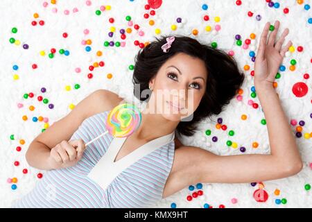 Young Latina woman laying on ruffled cloud like floor between colorful bubblegum balls holding a lollipop