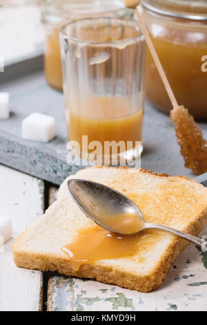 Toast with homemade caramel sauce for breakfast, served with spoon, jars and sugar cubes on gray tray over old wooden table Stock Photo