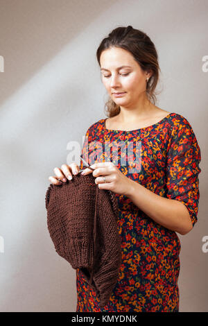 Close-up of a dark-haired pregnant woman in a summer dress in a floral print knits with natural shertail threads a brown sweater on a gray wall backgr Stock Photo