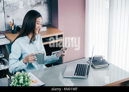 Asian businesswoman take a coffee break after working at laptop computer on desk with smiling face,Happy office life concept,working woman at modern h Stock Photo