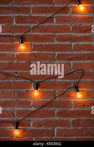 Brown brick wall with glowing orange lamp lights. Many bright electric bulbs on black wire. Vertical color photography. Stock Photo