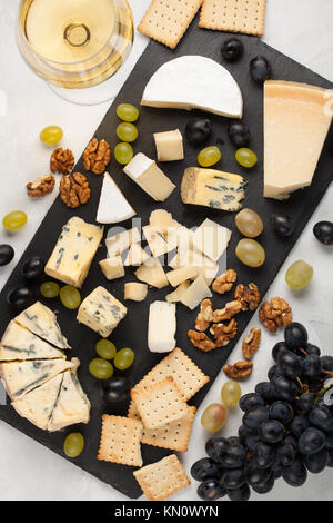Assorted cheeses with white grapes, walnuts, crackers and white wine on a stone Board. Food for a romantic date on a light background. Top view. Stock Photo