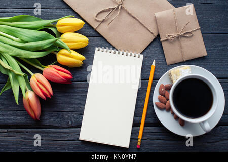 Notepad with pencil next to the tulips, coffee, and envelopes on wooden background. View from above with copy space. Stock Photo