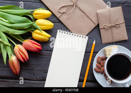Notepad with pencil next to the tulips, coffee, and envelopes on wooden background. View from above with copy space. Stock Photo