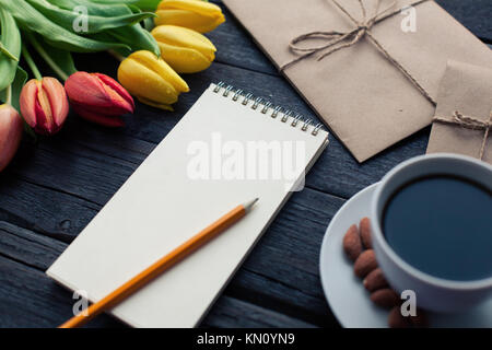Notepad with pencil next to the tulips, coffee, and envelopes on wooden background. View from above with copy space. Stock Photo