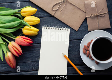 Notepad with pencil next to the tulips, coffee, and envelopes on wooden background. View from above with copy space. Stock Photo