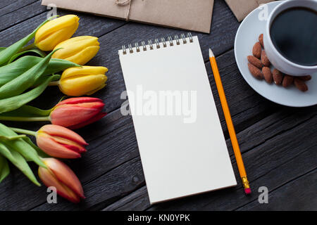 Notepad with pencil next to the tulips, coffee, and envelopes on wooden background. View from above with copy space. Stock Photo