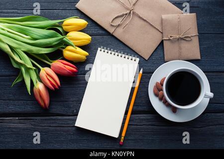 Notepad with pencil next to the tulips, coffee, and envelopes on wooden background. View from above with copy space. Stock Photo