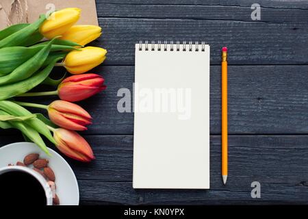 Notepad with pencil next to the tulips, coffee, and envelopes on wooden background. View from above with copy space. Stock Photo