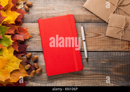 Red notebook with pen and paper envelopes on old wooden table. Mixed maple autumn leaves and acorns next to a closed notebook. Stock Photo