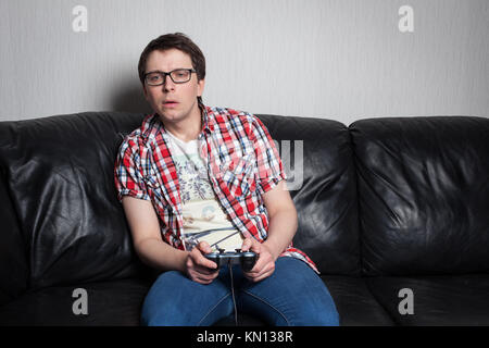 Young guy with glasses and red shirt playing video games on the joystick, sitting on a black leather sofa. Stock Photo