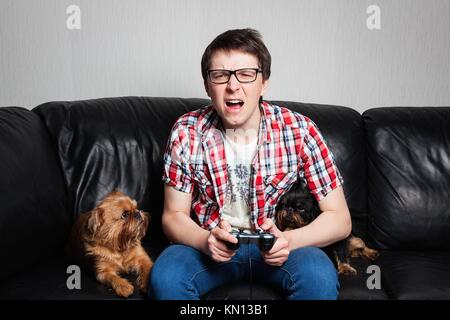 A young man in a red shirt and blue jeans sits at home and plays video games together with their dog. Screaming boy in front of tv trying to fight wit Stock Photo
