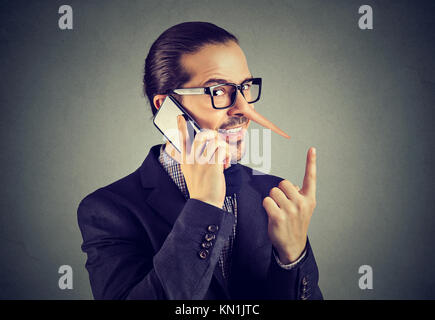Sly liar business man with long nose talking on mobile phone isolated on gray wall background. Stock Photo