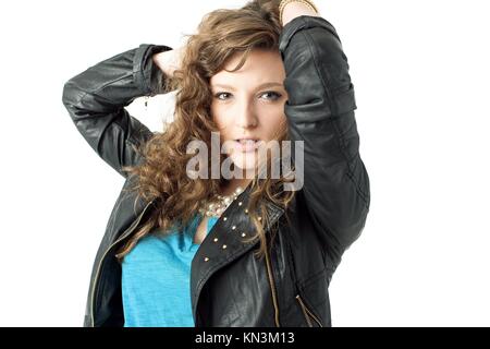 Young pretty woman in a blue shirt and black leather jacket has both arms up behind her head as she looks at the camera.