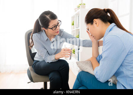 beautiful young girl visiting female psychologist frustration crying and doctor woman giving patient glass of water trying to calm her down. Stock Photo