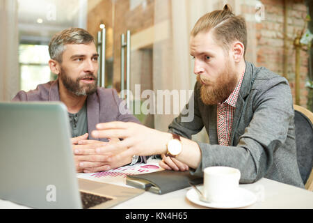 Portrait of two successful business men discussing work and using laptop during meeting in cafe on coffee break, focus on hipster businessman pointing Stock Photo