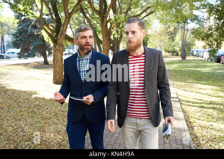 Portrait of two handsome modern businessmen discussing work walking on lane in green park, copy space Stock Photo