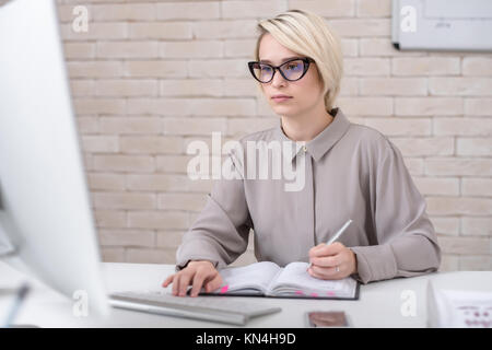 Portrait of blonde successful businesswoman using computer for work sitting at desk in modern office and making notes Stock Photo