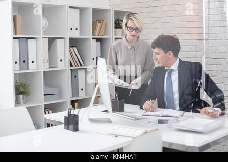 Portrait of boss sitting at desk in modern office behind glass wall with secretary handing him papers, copy space Stock Photo