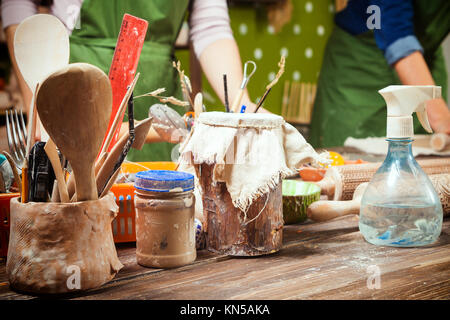 A close-up of the tools for the creativity of the potter (brushes, spoons, scissors) on a wooden table, in the background, two women mold clay from Ne Stock Photo