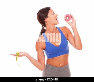 Fit woman in sport clothing holding measuring tape while biting a cake against white background.