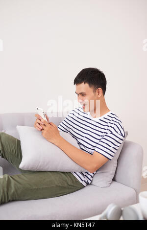 Young aisan man sitting relaxed on sofa with smartphone checking his smartphone messages Stock Photo