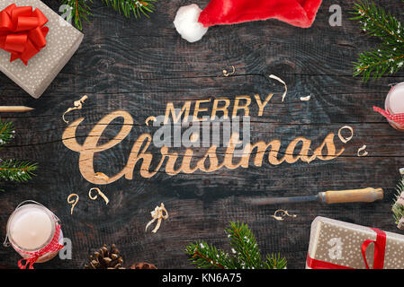 Merry Christmas greeting carved into a wooden surface. Christmas gifts, tree branches, chisel, candles, pinecones and Santa Claus hat beside. Stock Photo