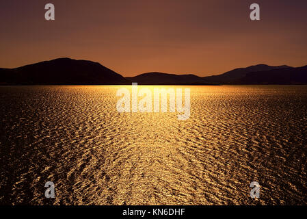 Dark bronze sunset with sun shimmering sun reflections on lake under clear bronze skies Stock Photo