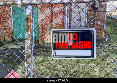 A No Trespassing sign on a chain link fence at an abandoned brick factory building with broken windows. Stock Photo