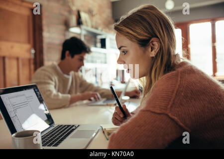 Woman looking at laptop for making notes with her boyfriend working on laptop in background. Couple sitting at table and working from home. Stock Photo