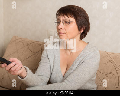 Elderly woman in glasses sitting on couch with remote control in hand Stock Photo