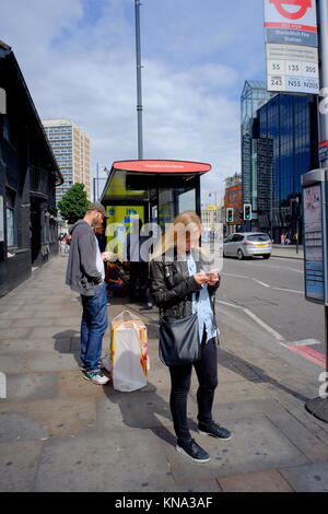 People using smartphones whilst waiting at bus stop in London, England, UK Stock Photo