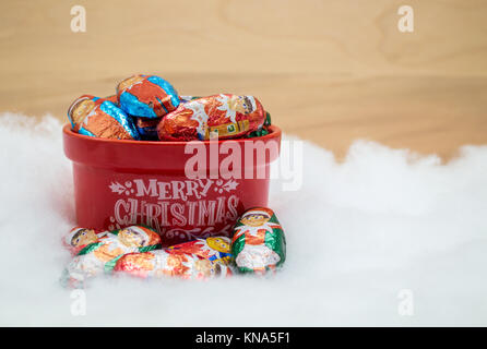 red Merry Christmas bowl filled with chocolate elves wrapped in colorful foil against a wood wall Stock Photo