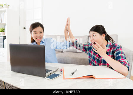 cheerful pretty female students using mobile computer doing homework at home and finished making school report clapping for each other celebrating. Stock Photo