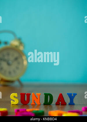 Colorful wooden word SUNDAY on wooden table and vintage alarm clock and background is powder blue. English alphabet made of wooden letter color. Stock Photo