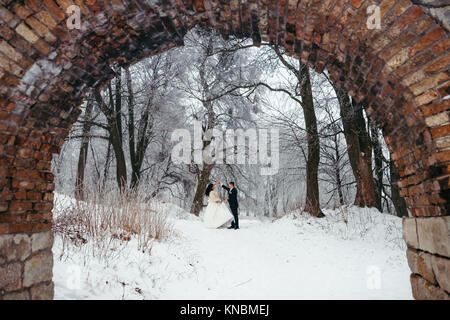 Dancing happy newlywed couple in the snowy forest. View through the old stone arch. Stock Photo