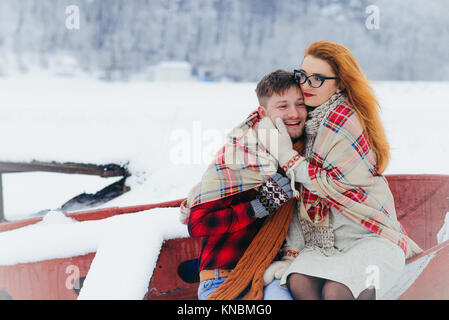 Sensitive half-length portrait of the beautiful young woman stroking the cheek of the handsome man. They are wrapped in the knitted plaid while sitting on the boat during the snowfall. Stock Photo