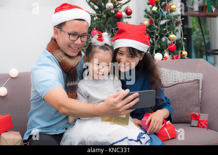 Happy family Asia selfie with mobile at sofa,father and mother wear santa claus hat give Christmas gift box to child at house xmas party,Holiday celeb