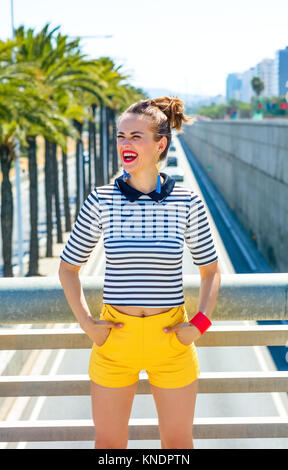 Nosing around, having fun. smiling trendy woman in yellow shorts and stripy shirt against highway in the city looking into the distance Stock Photo