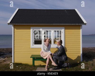 Close up of attractive young couple on the beach in front of cute yellow beach hut on a windy winter's day looking into each others eyes Stock Photo