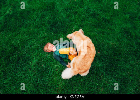 Overhead view of a boy lying on the grass playing with his golden retriever dog Stock Photo