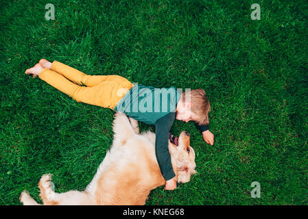 Overhead view of a boy lying on the grass playing with his golden retriever dog Stock Photo