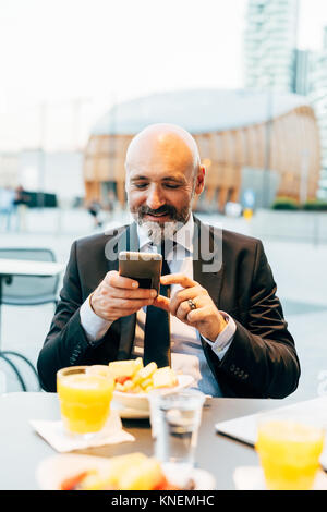 Mature businessman sitting outdoors at cafe, using smartphone Stock Photo
