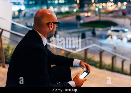 Mature businessman outdoors at night, sitting on steps, holding smartphone Stock Photo