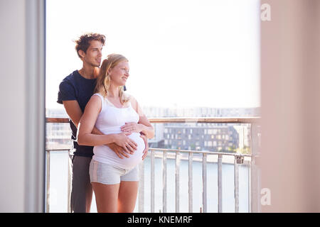 Pregnant couple looking out from waterfront apartment balcony Stock Photo